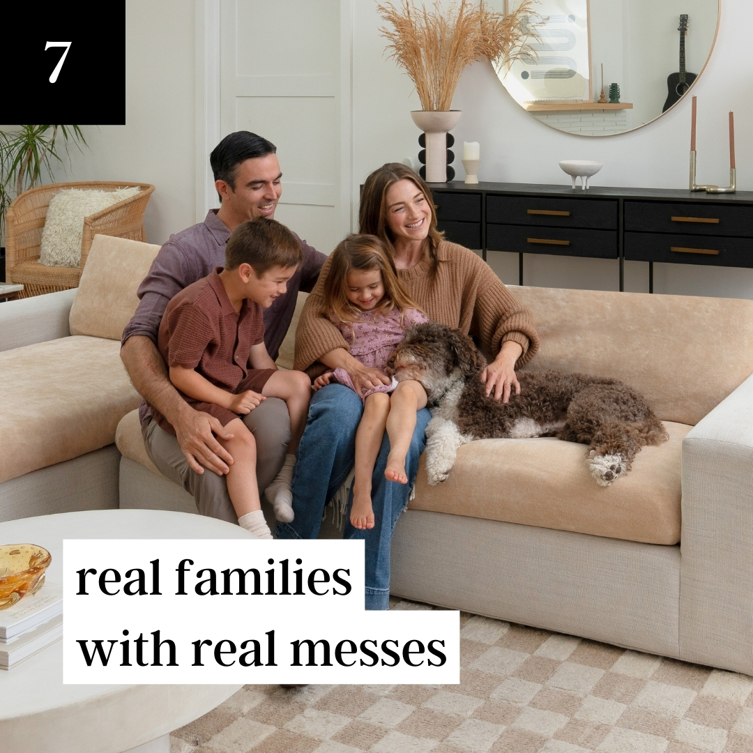 A family of four sits smiling on a beige sectional sofa with their dog in a living room.