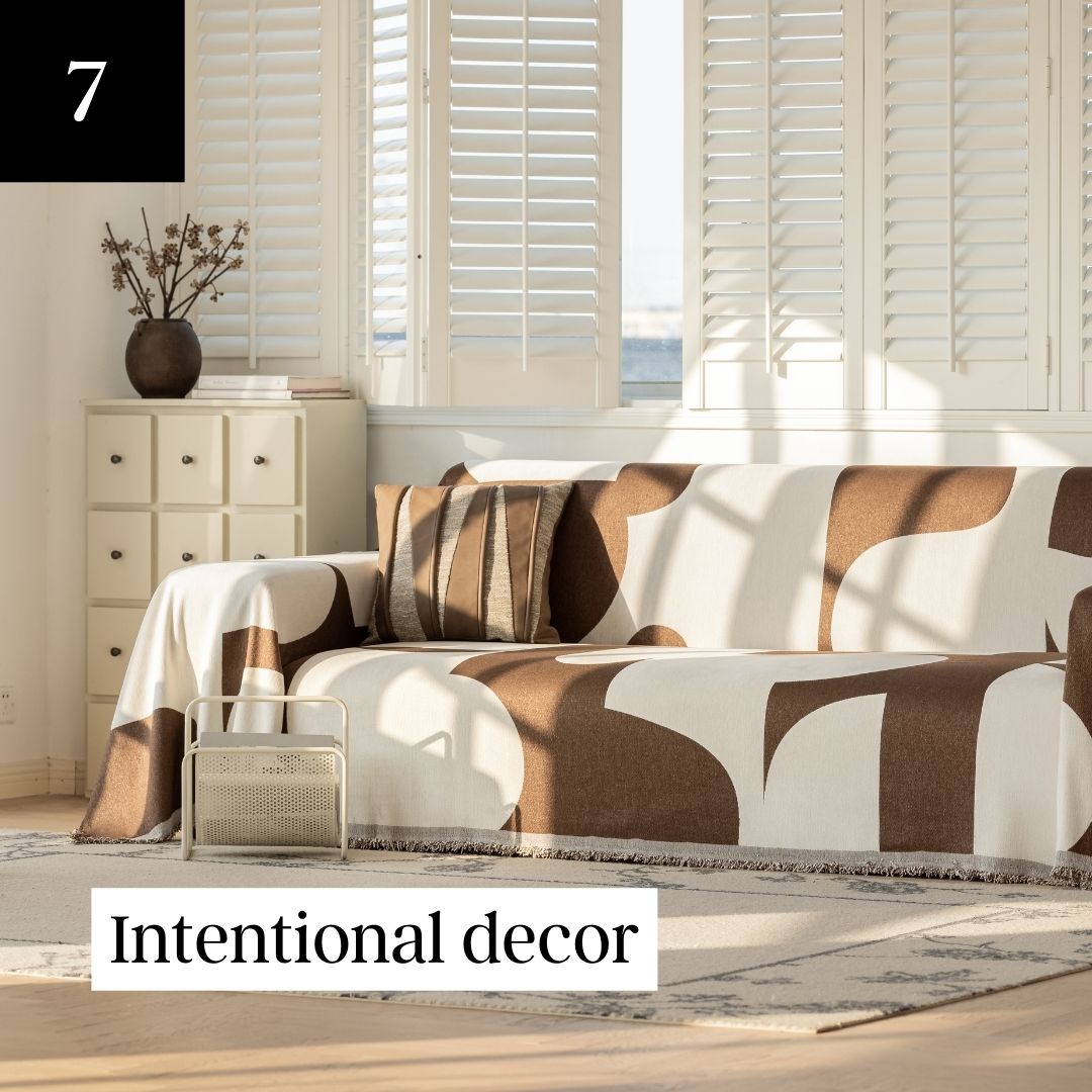 A sunlit room with a sofa covered by a brown and white patterned throw, next to a window with shutters.