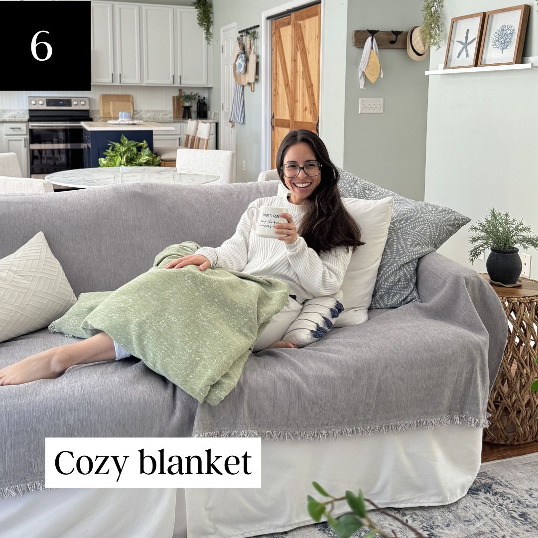 Person with glasses smiling, wrapped in a green blanket on a couch, holding a mug.