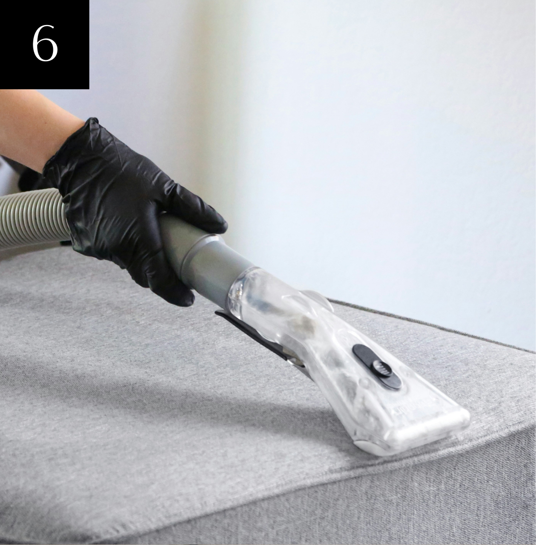 Gloved hand using a vacuum cleaner on a gray upholstered surface.