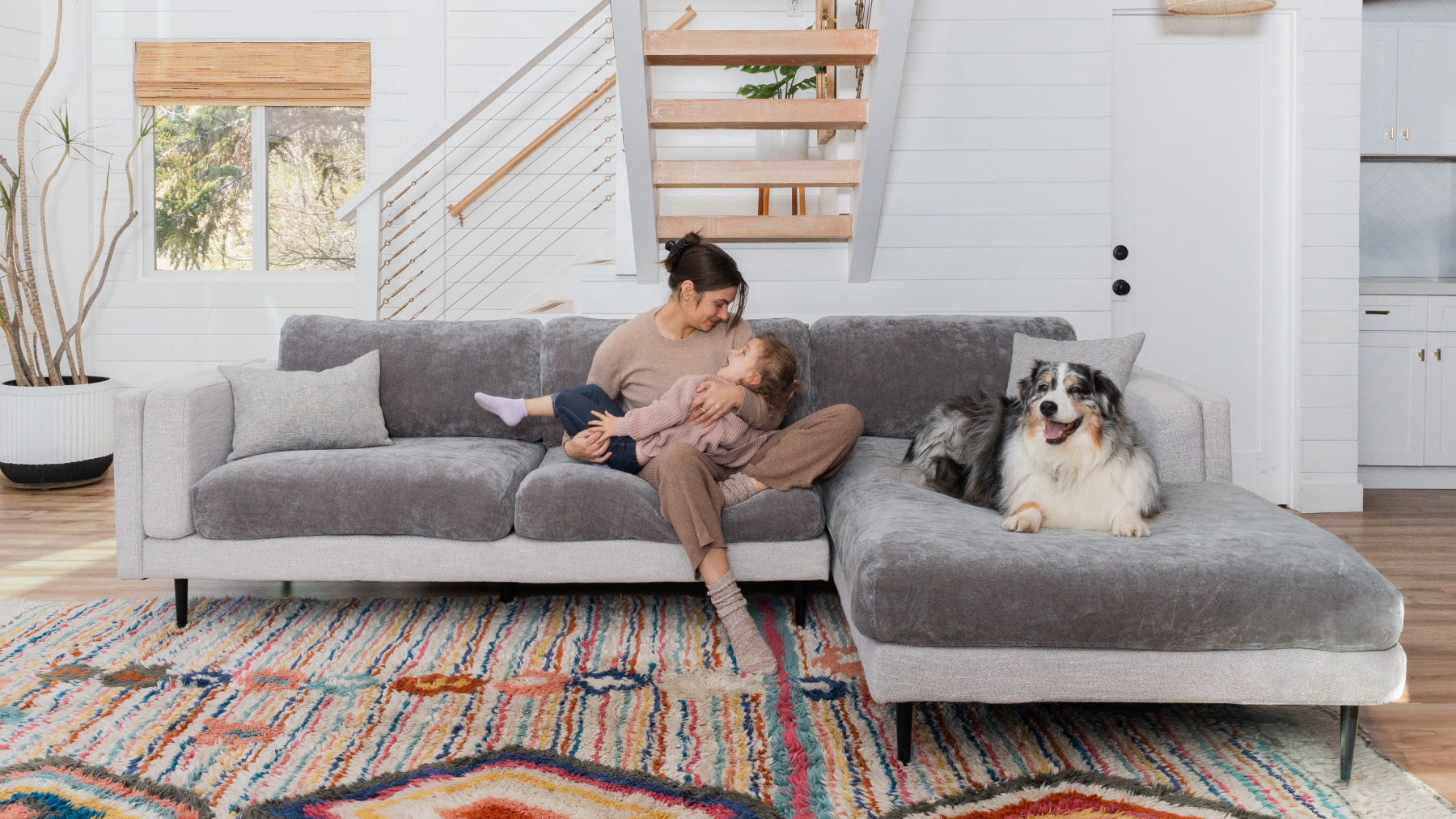 Woman and child sitting on a gray sofa with a dog nearby in a living room.