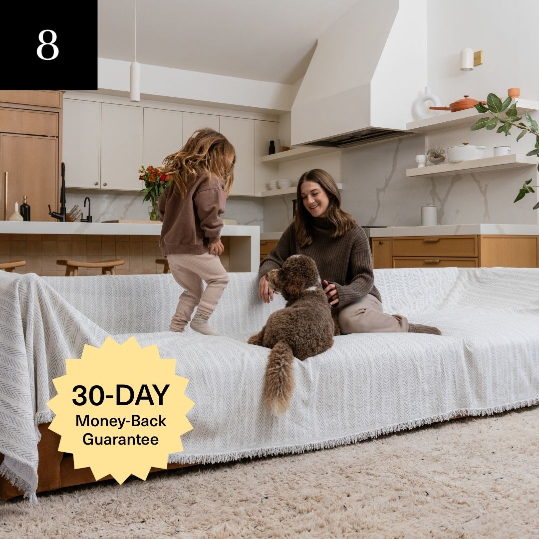 A woman, a child, and a dog sit on a couch covered with a white, patterned blanket.