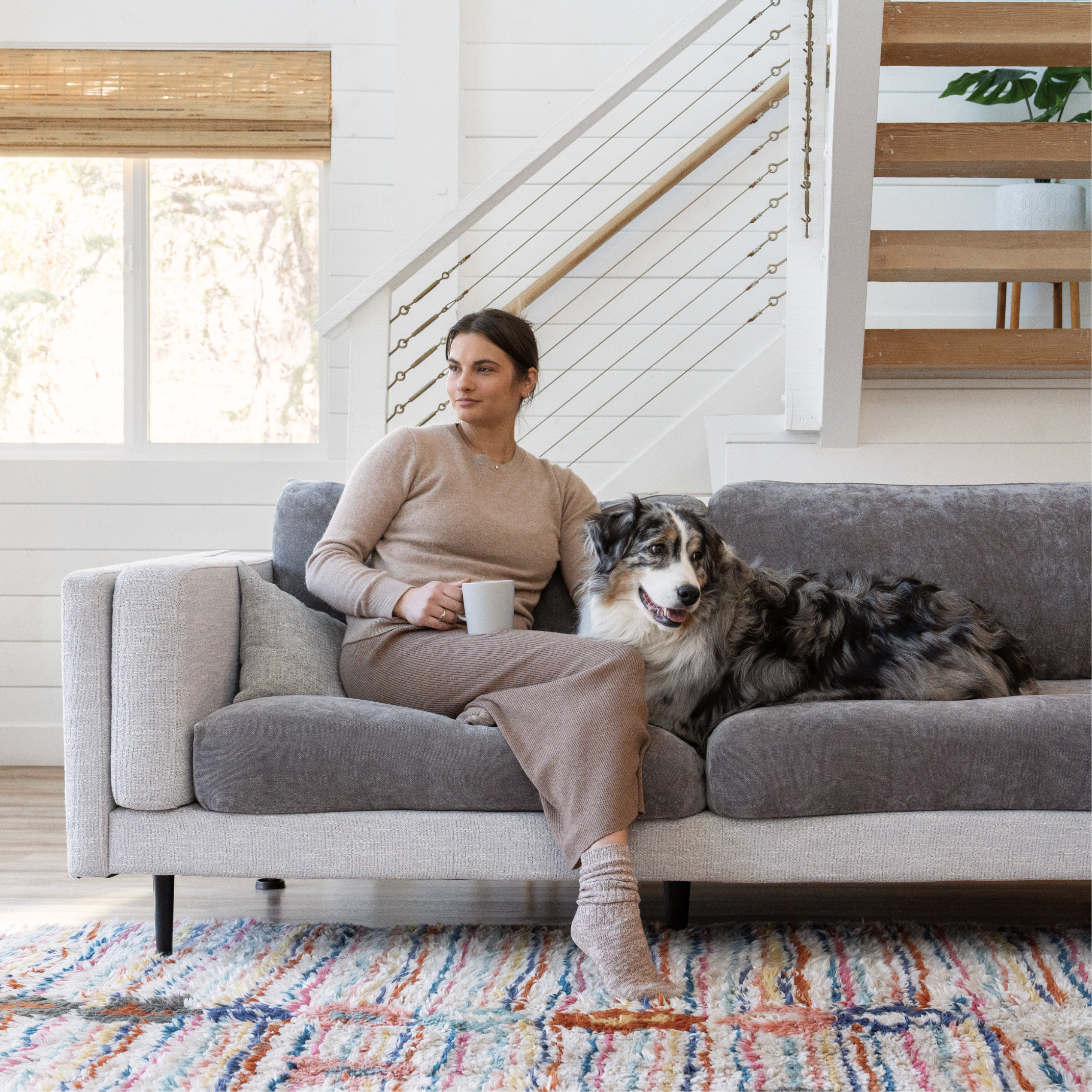 Woman with a mug and a dog on a gray sofa in a living room.