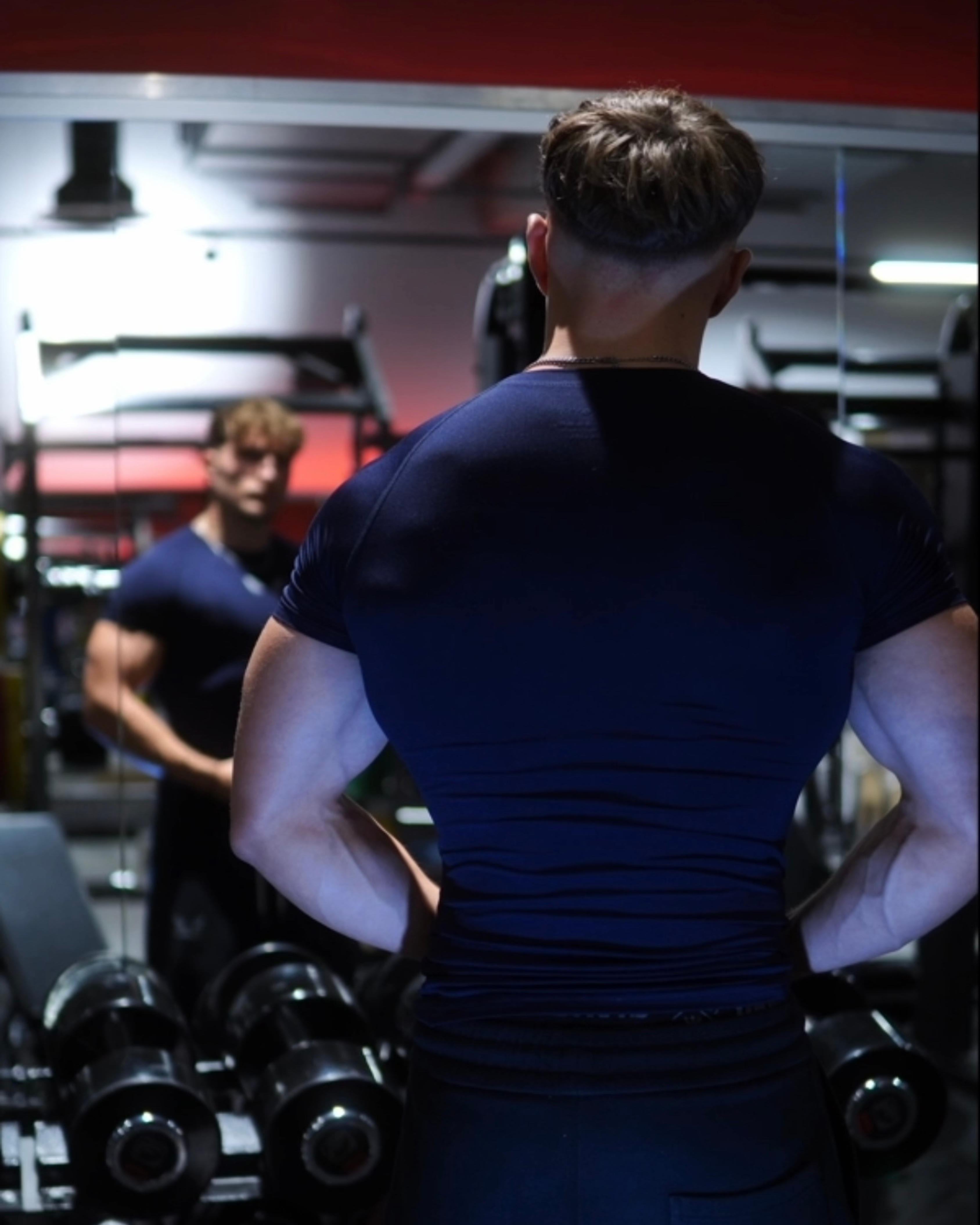 Person in gym looking at reflection in mirror with weights nearby.