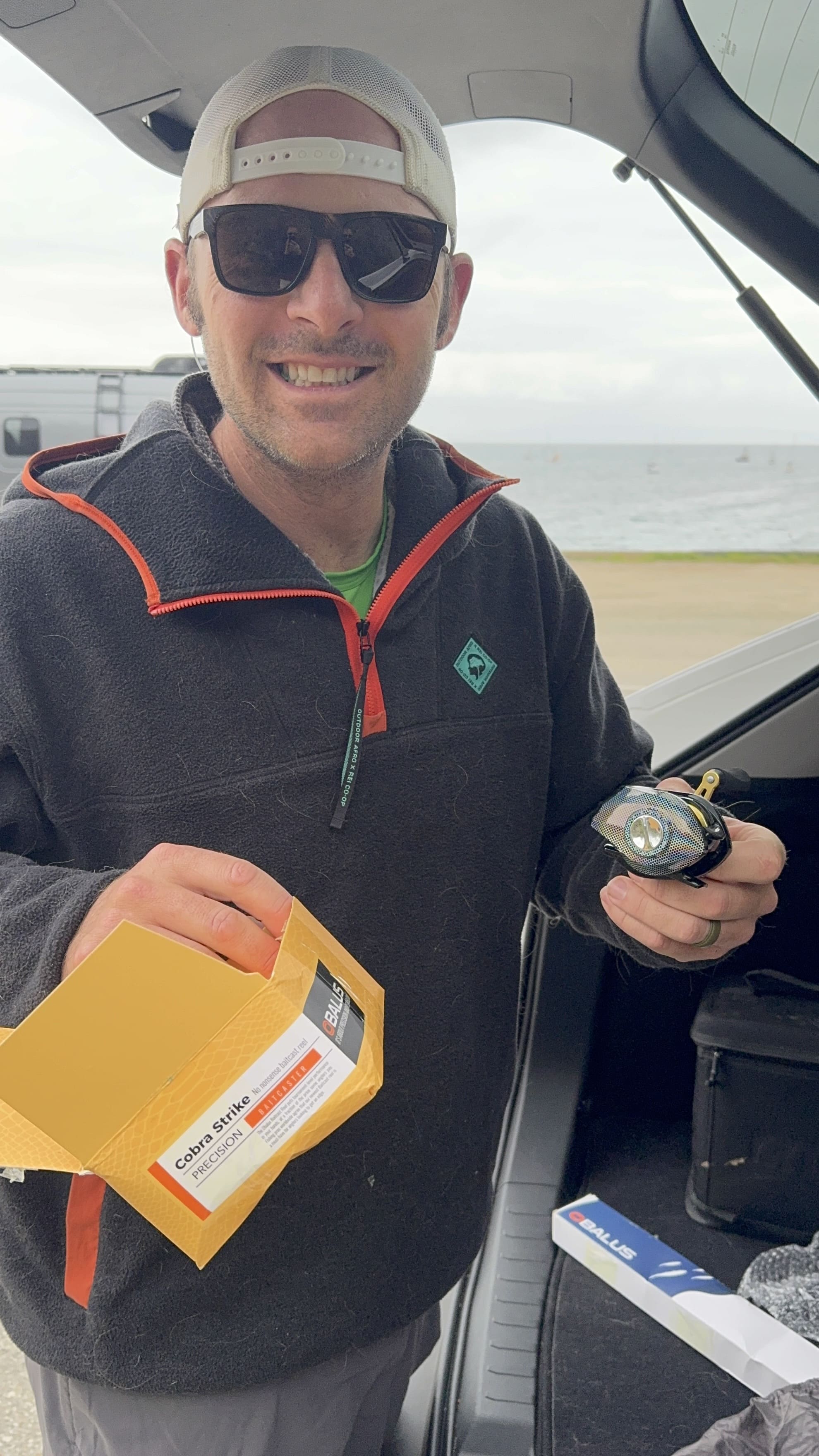 Man holding a package and an object, standing near a car by the water.