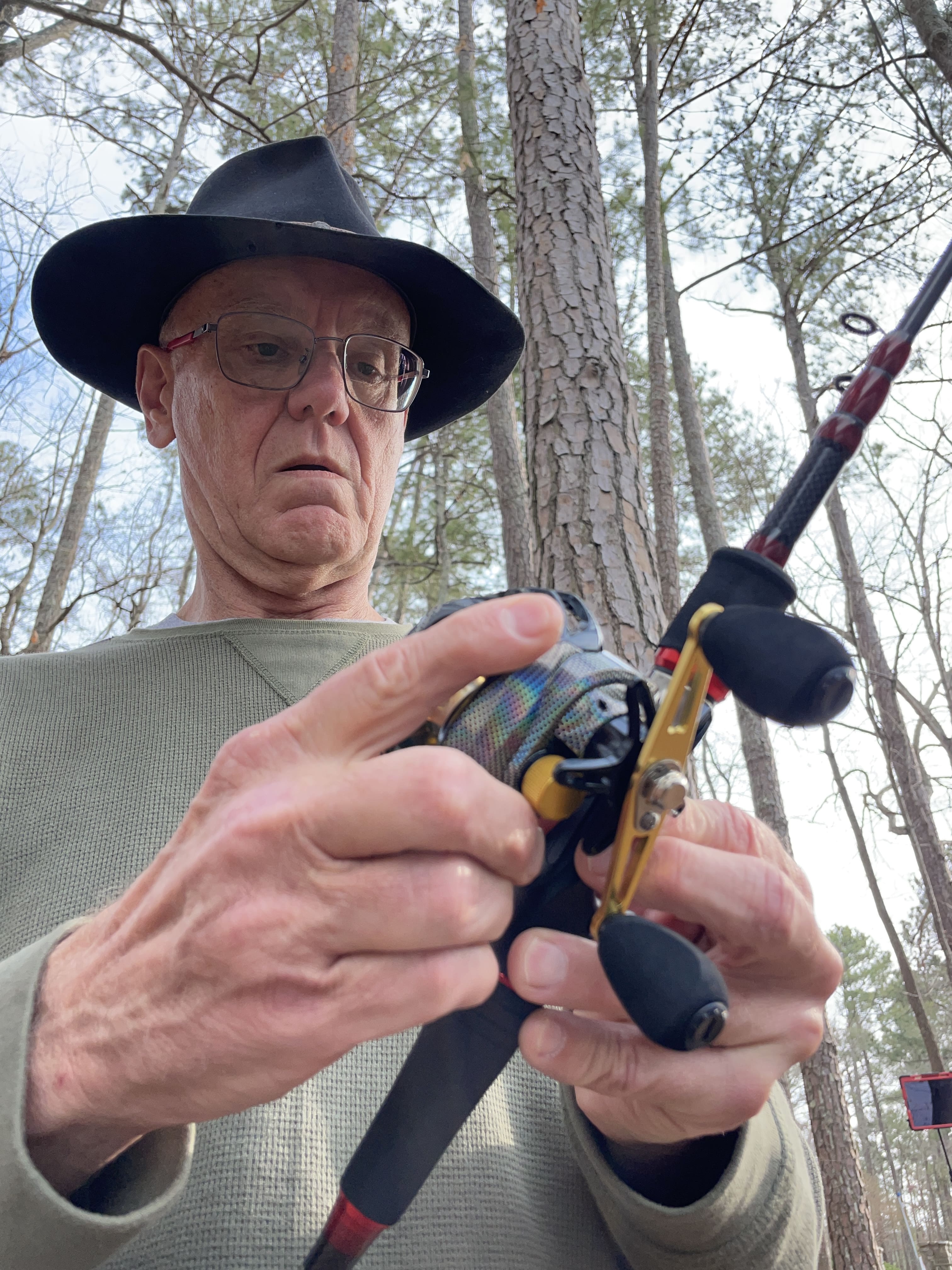 Man adjusting a fishing rod in a forest wearing a black hat and glasses.