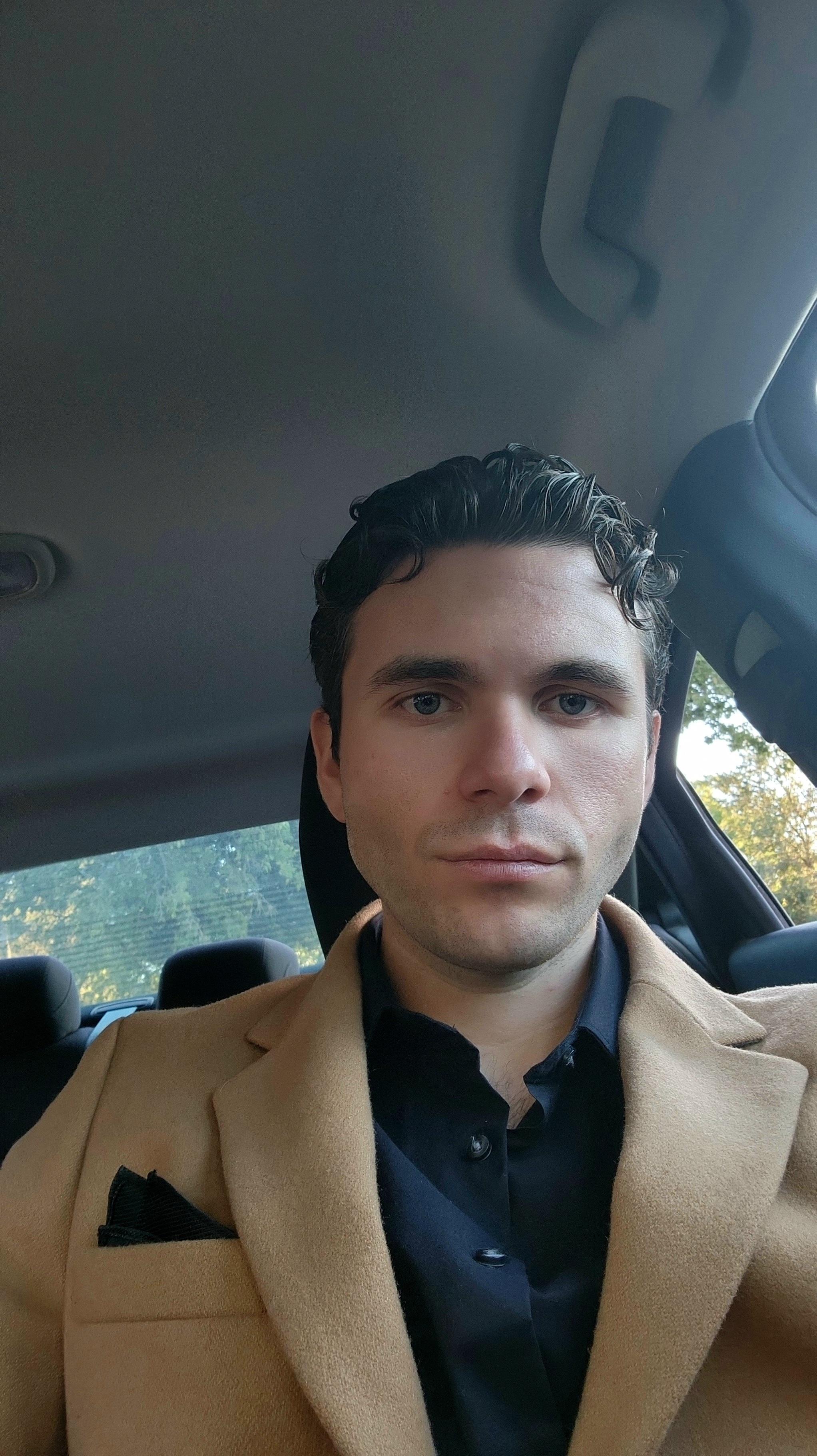Man in a tan coat sitting inside a car.