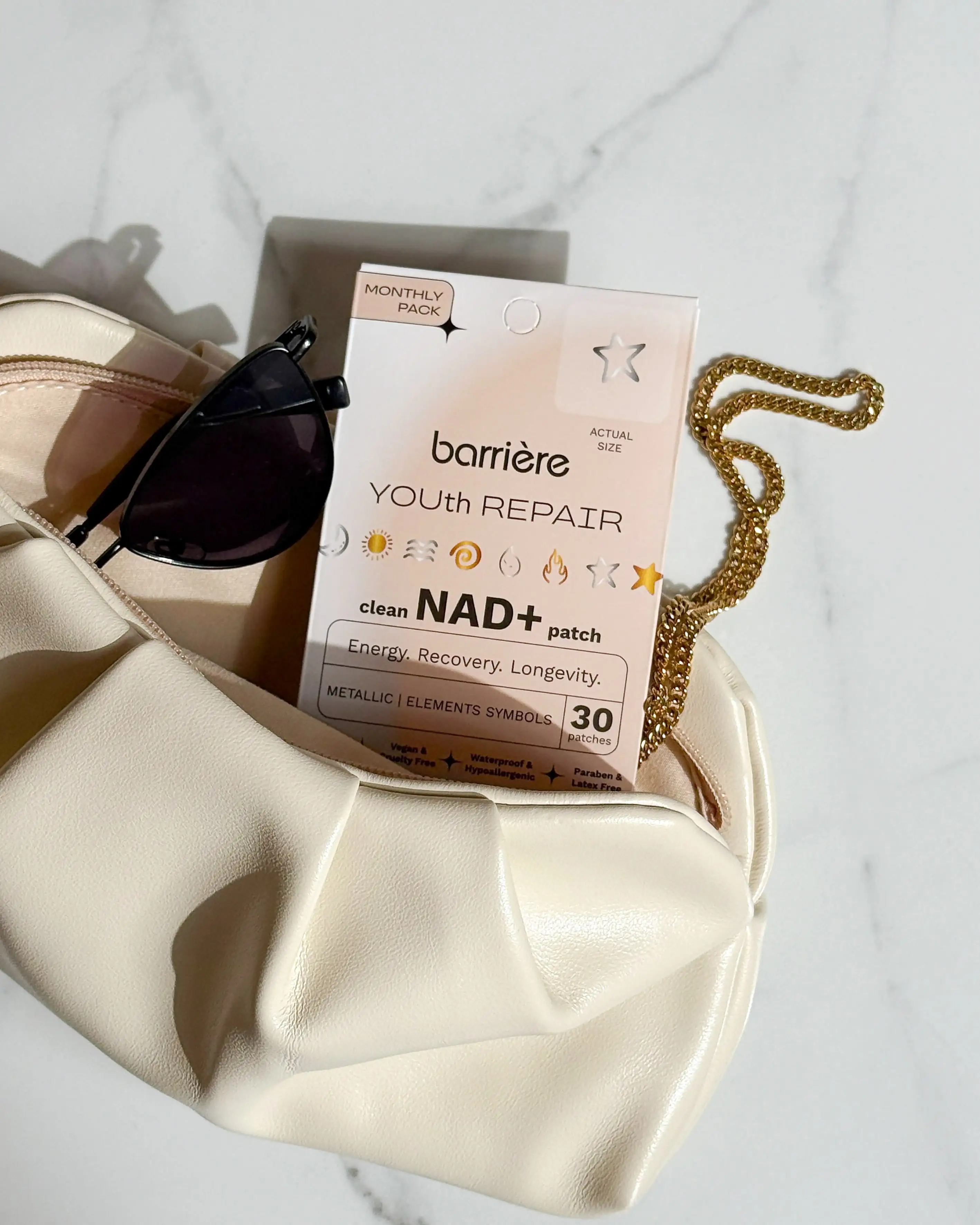 A box of Barrière Youth Repair patches and sunglasses sit inside a white handbag on a marble surface.