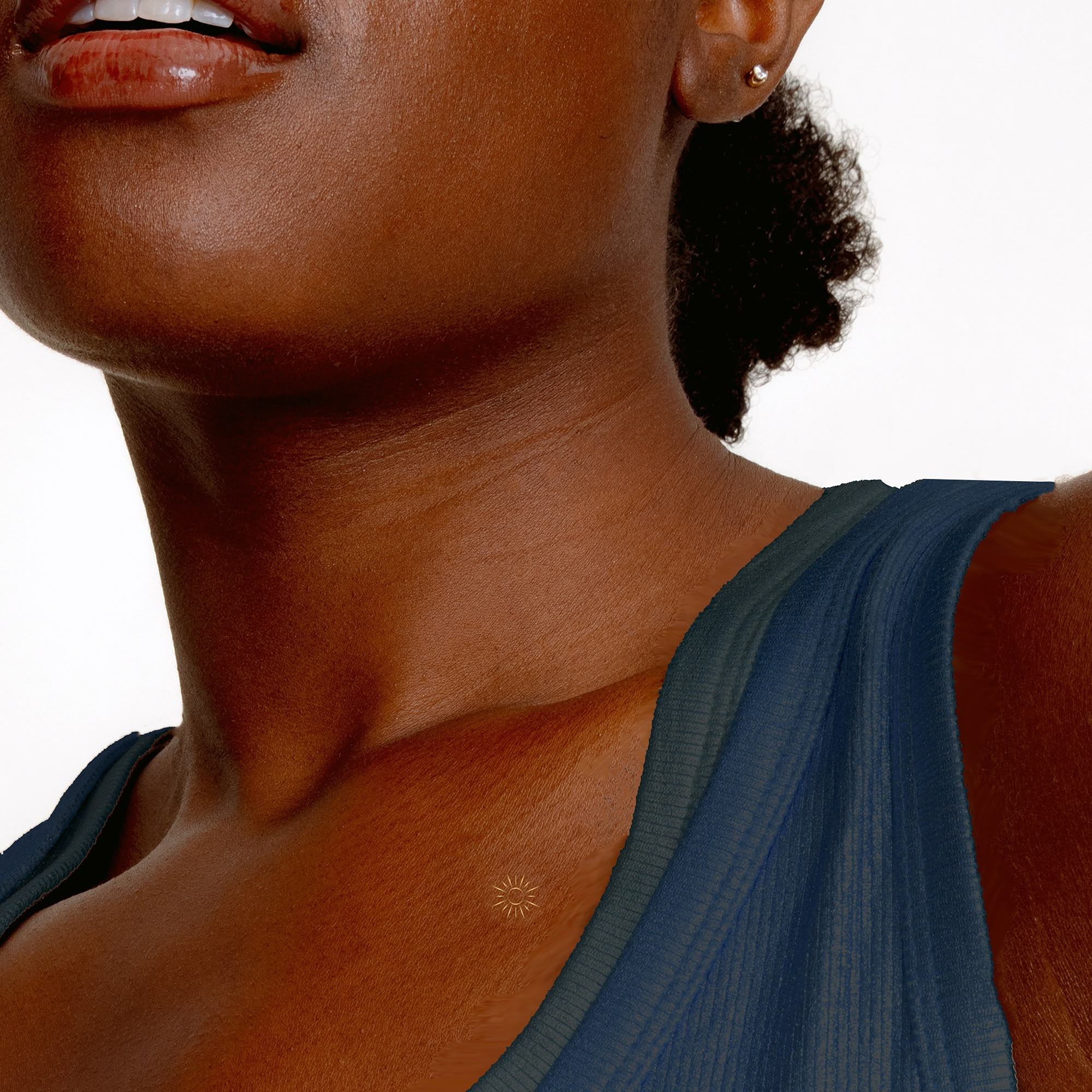 A close-up of a Black woman's neck and shoulder, wearing a blue top, with a small sun tattoo.