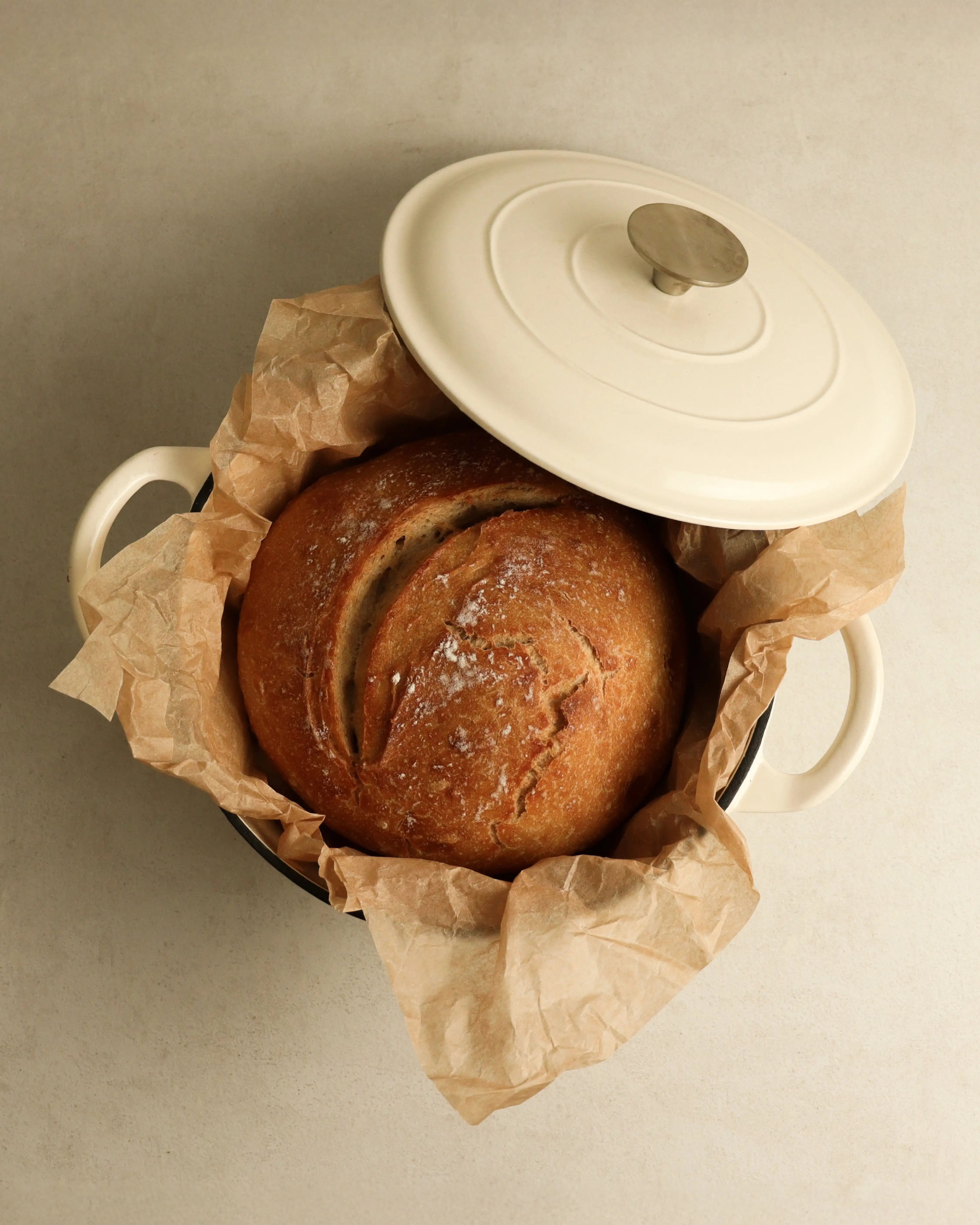 A freshly baked round loaf of bread sits on parchment paper inside a white dutch oven.