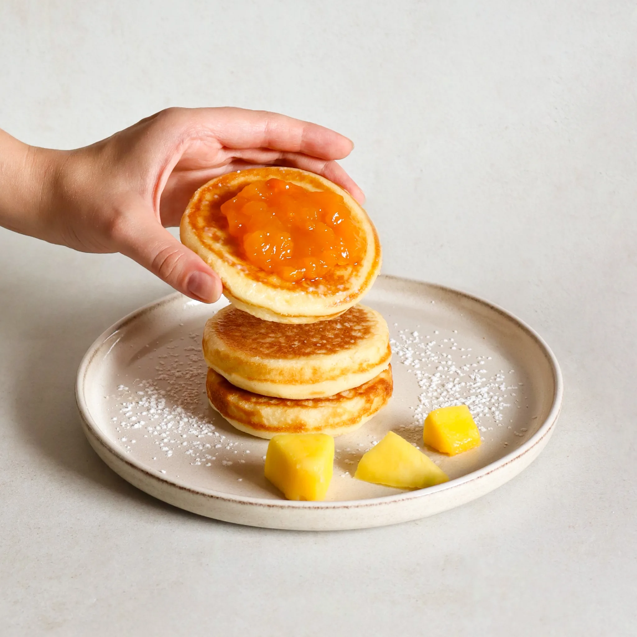 A hand places a pancake with orange jam onto a stack on a plate with mango.