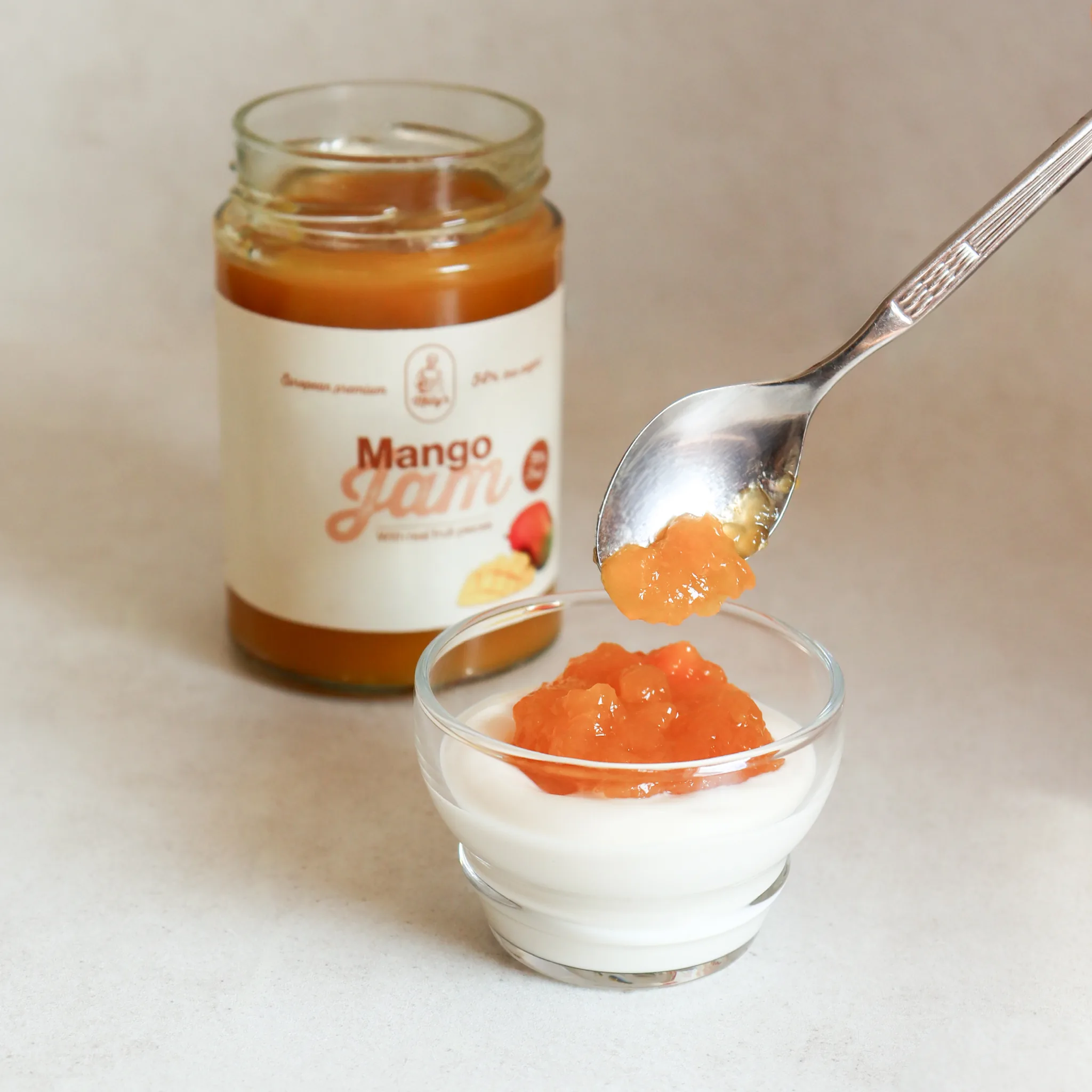 A spoonful of mango jam being added to a glass bowl of yogurt, with the jar behind.