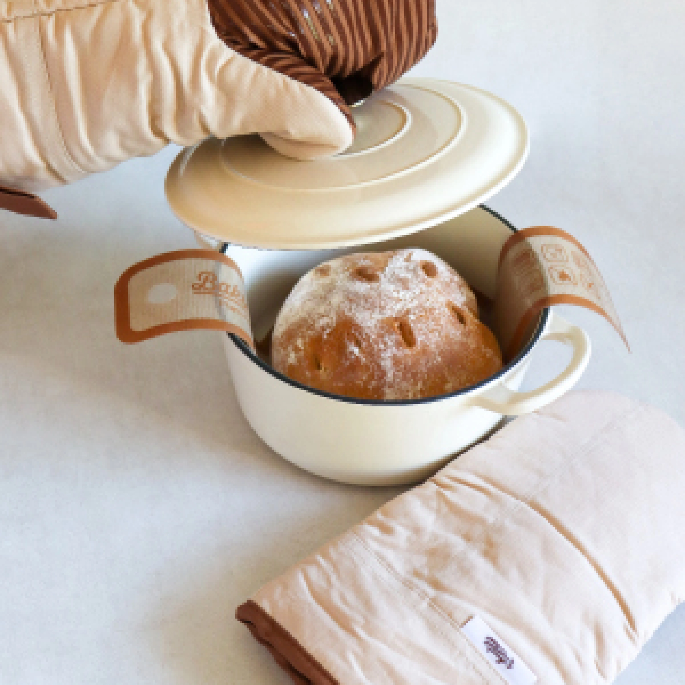 A person wearing oven mitts lifts the lid from a pot revealing a freshly baked loaf of bread.