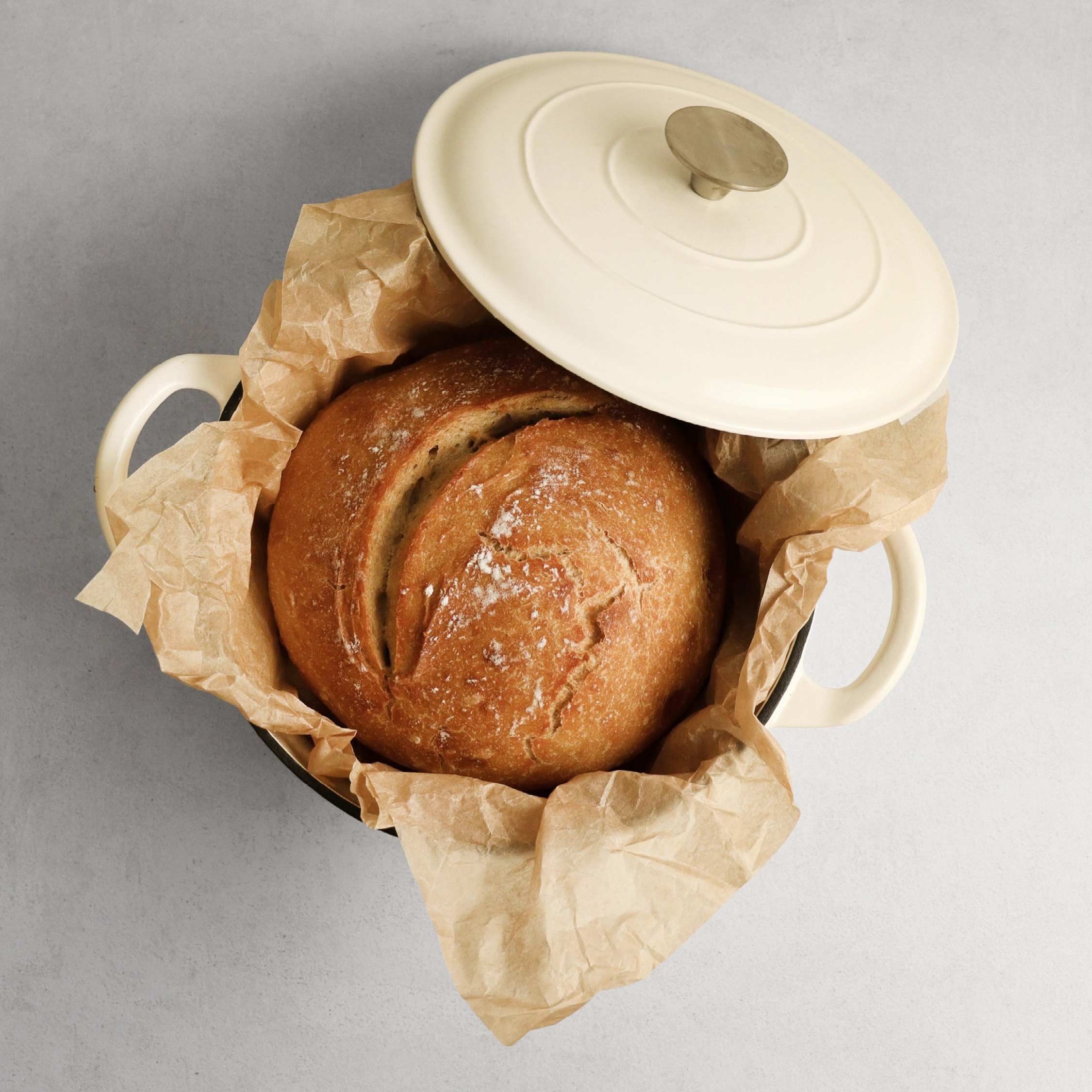 An overhead view of a round loaf of bread on parchment paper in a cream-colored dutch oven.