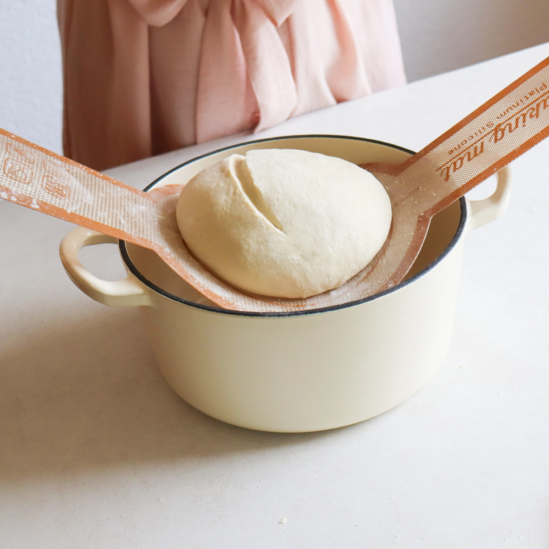 A person lowers a scored ball of dough into a pot using a silicone baking mat sling.
