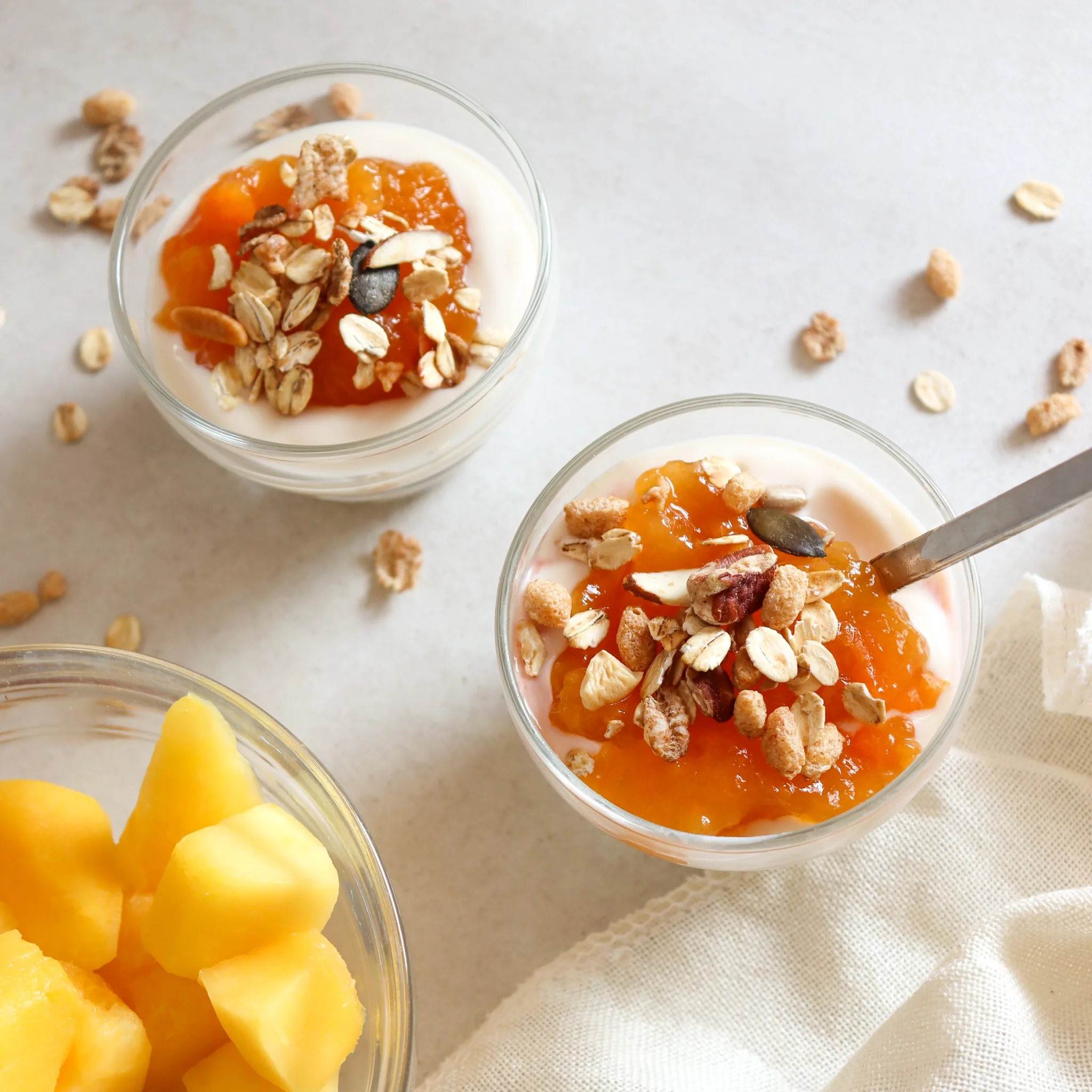 Two glass bowls of yogurt topped with orange jam and granola, with a side of fruit.