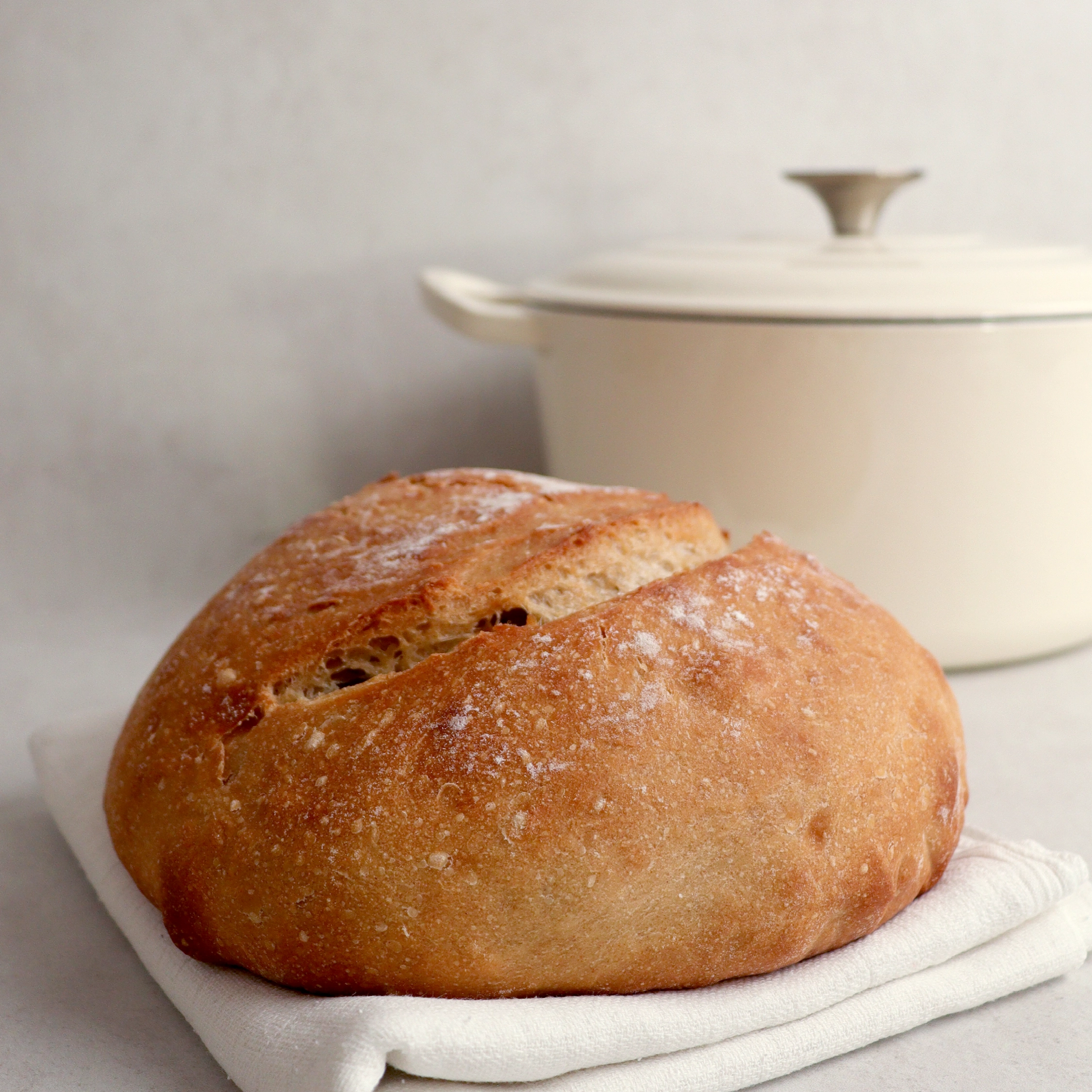 A round loaf of crusty bread sits on a white cloth in front of a white Dutch oven.