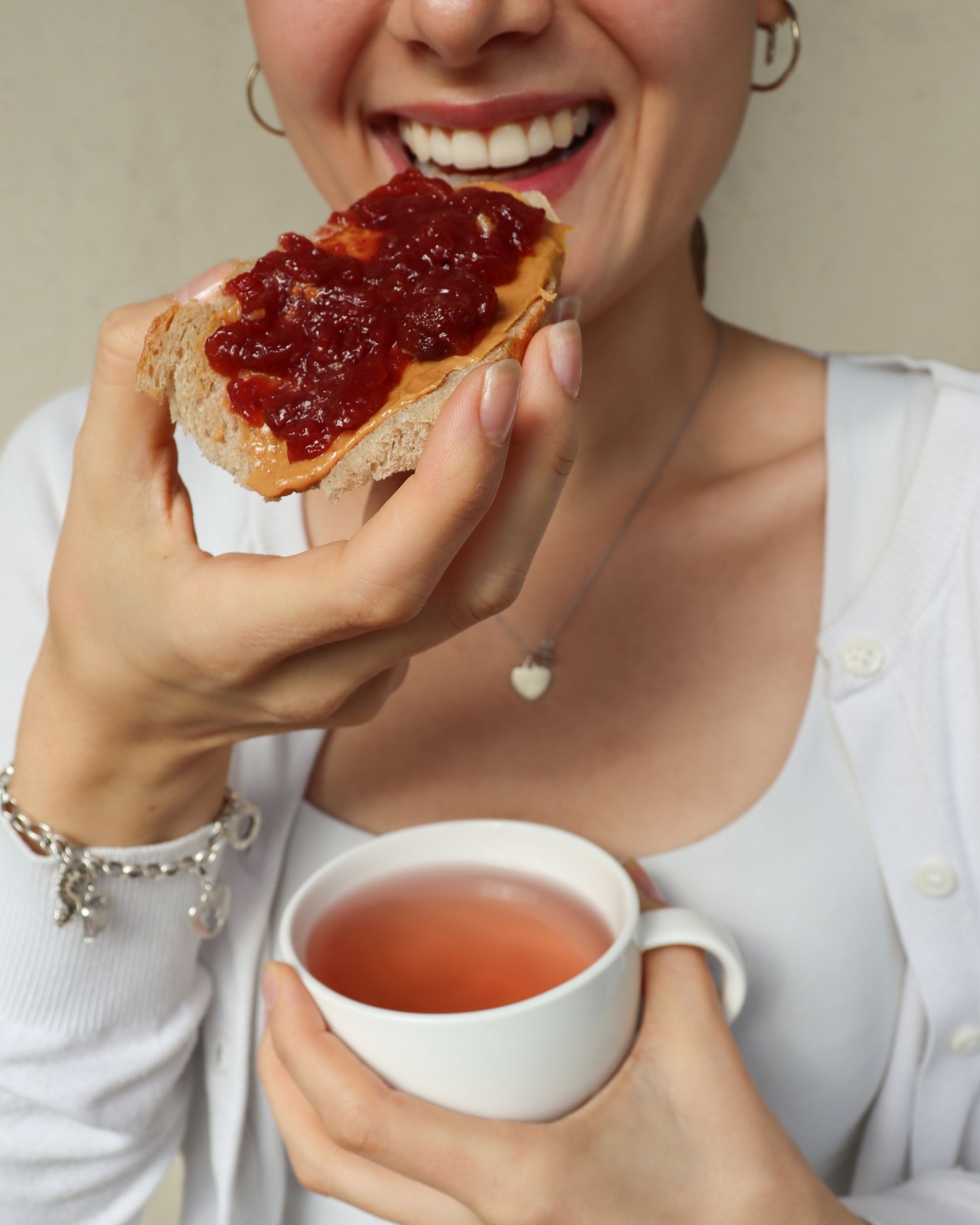 A smiling woman eating toast with peanut butter and jelly while holding a white mug of tea.
