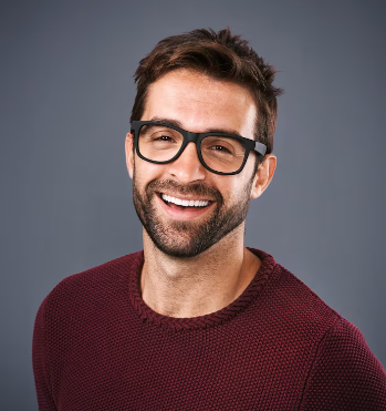 A smiling man with a beard and black-rimmed glasses wearing a maroon sweater against a grey background.