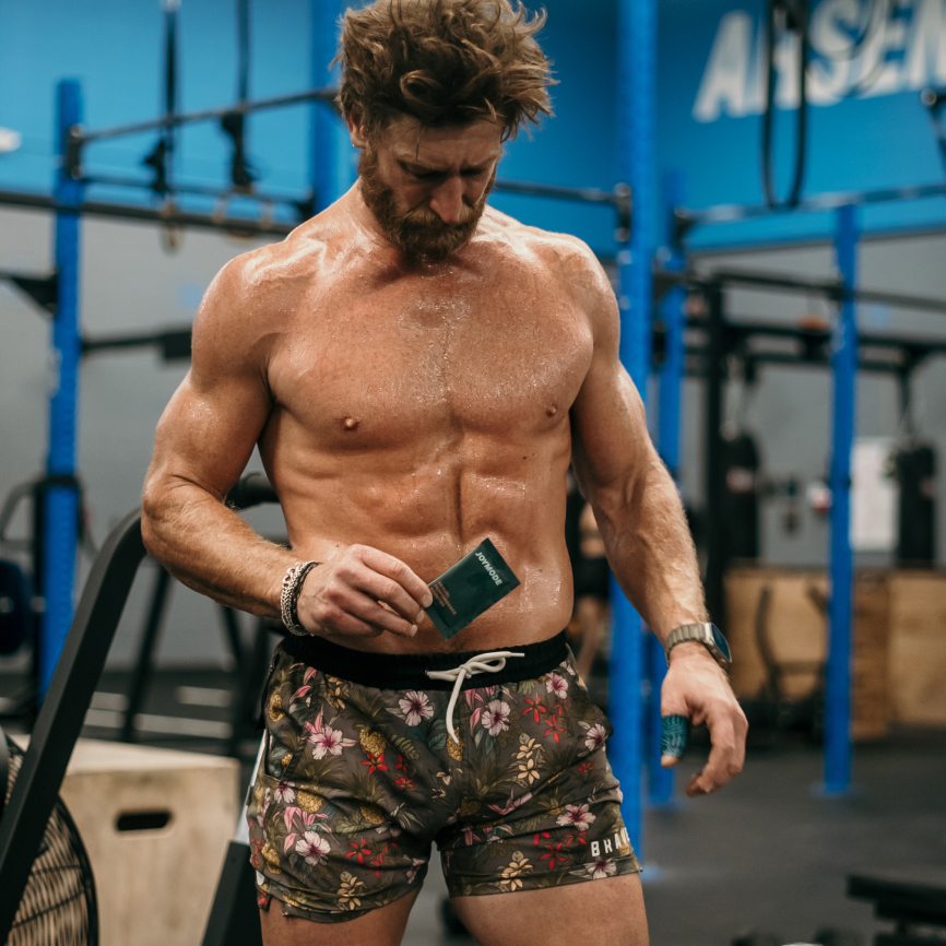Muscular man in gym holding a small packet, wearing floral shorts.