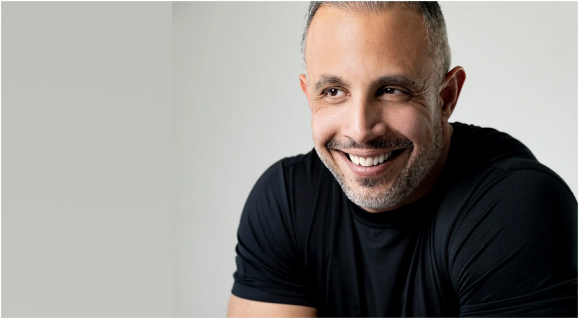 A smiling man with short, graying hair and a beard, wearing a black t-shirt against a plain background.