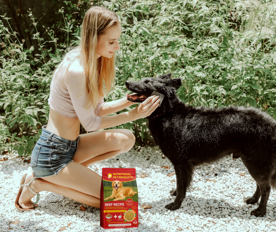 Woman kneeling, petting a black dog; dog food bag in the foreground, plants in the background.