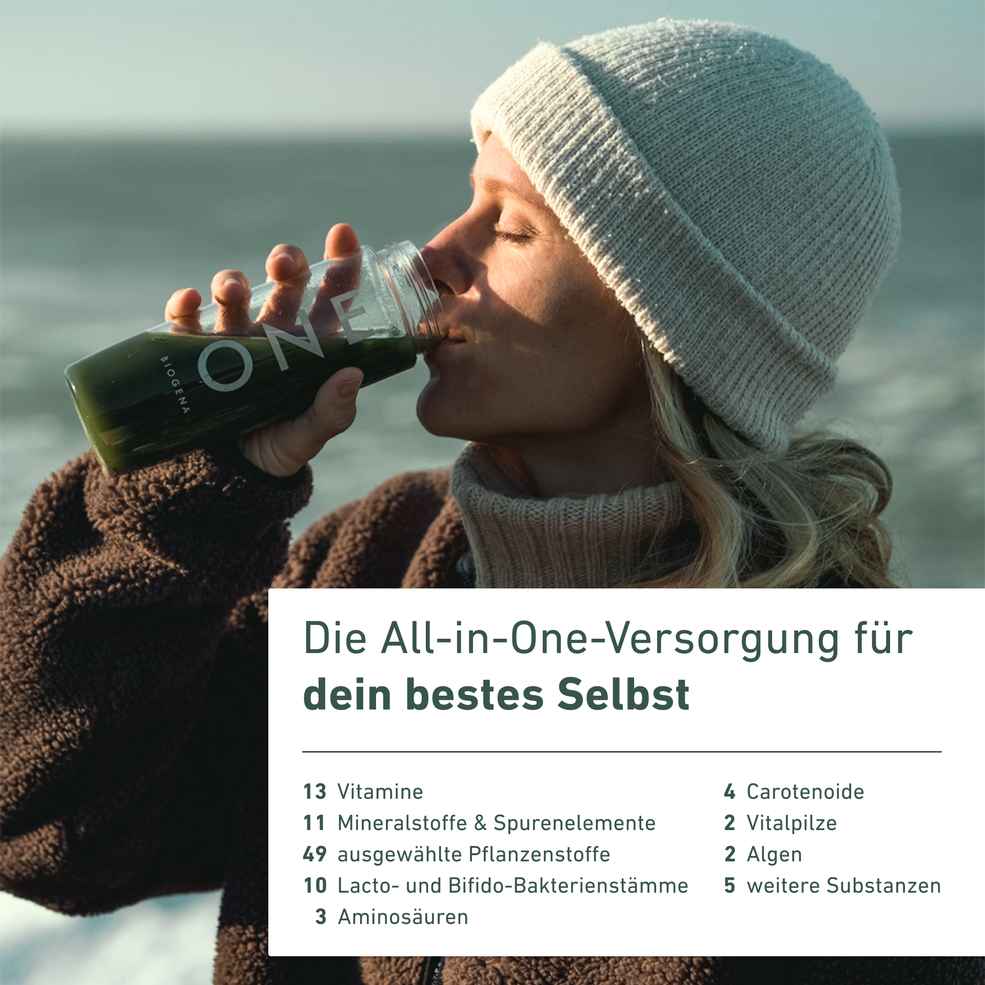 Person in a beanie drinking from a bottle labeled 'ONE' by the sea.