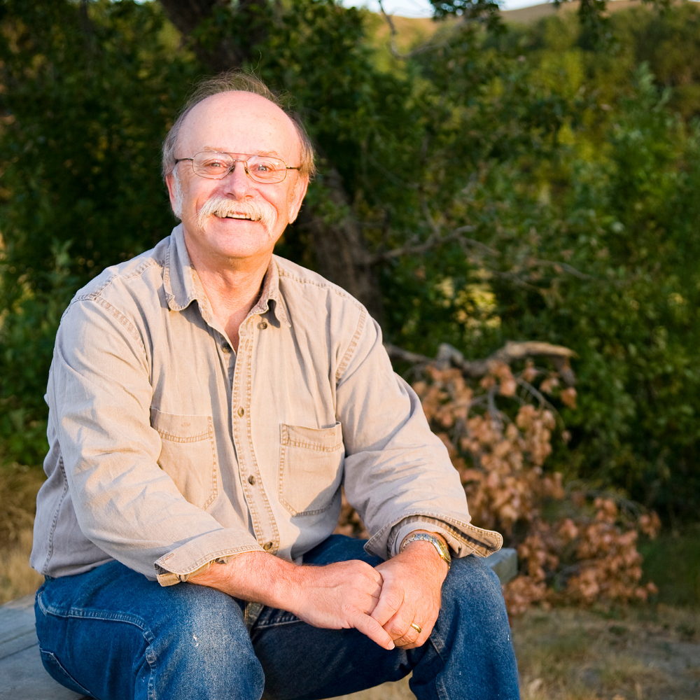Smiling man with glasses in a blue shirt, outdoors with blurred background.