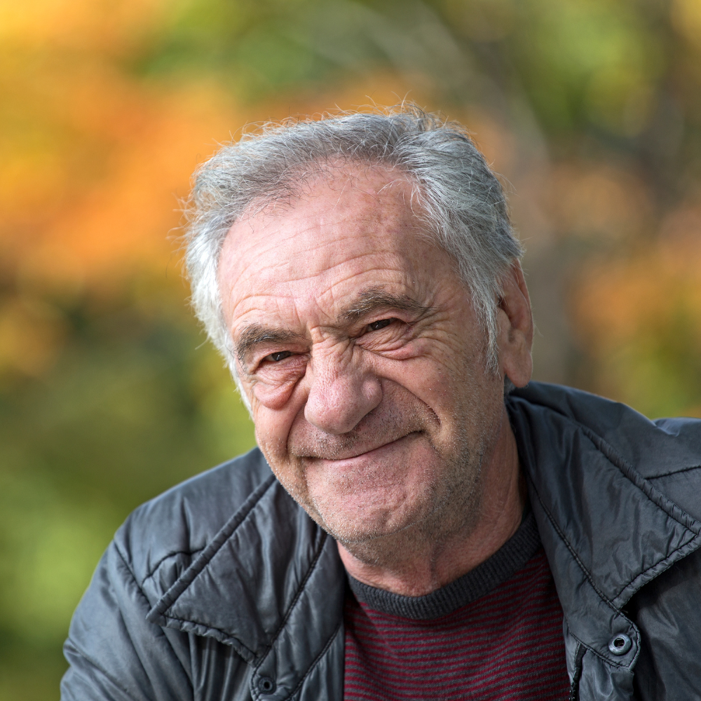 Smiling older man with gray hair in a jacket, outdoor setting with blurred autumn colors.