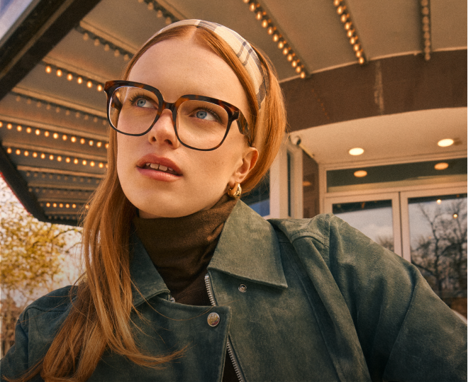 A woman with red hair wearing tortoiseshell glasses and a green jacket stands under a lighted marquee.