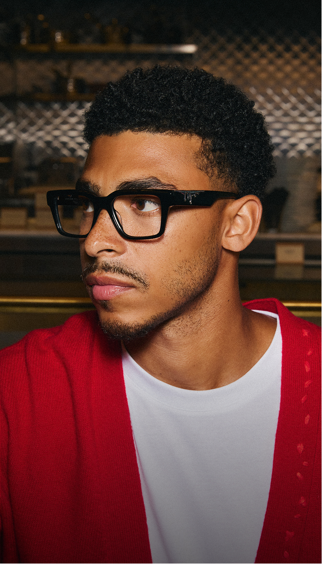 A young man wearing a red cardigan and thick black-framed glasses looks off to the side.