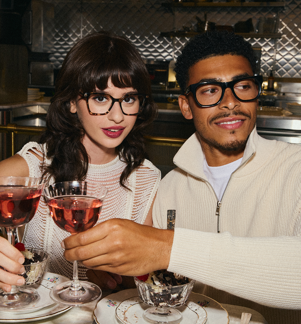 A man and a woman wearing glasses sit at a restaurant table, toasting with drinks and dessert.