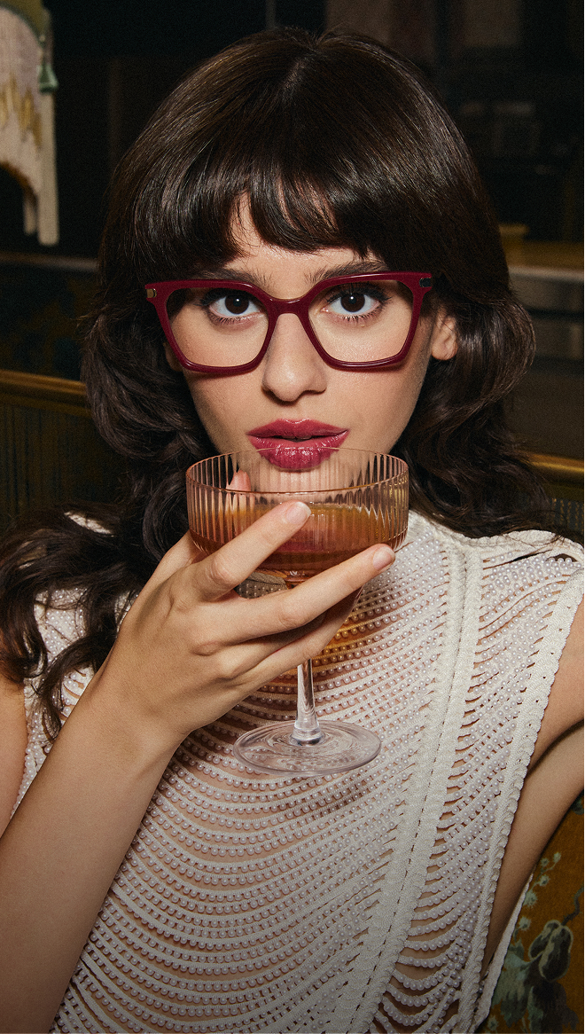 A woman with dark hair and bangs wearing magenta glasses holds a cocktail glass and looks at the camera.