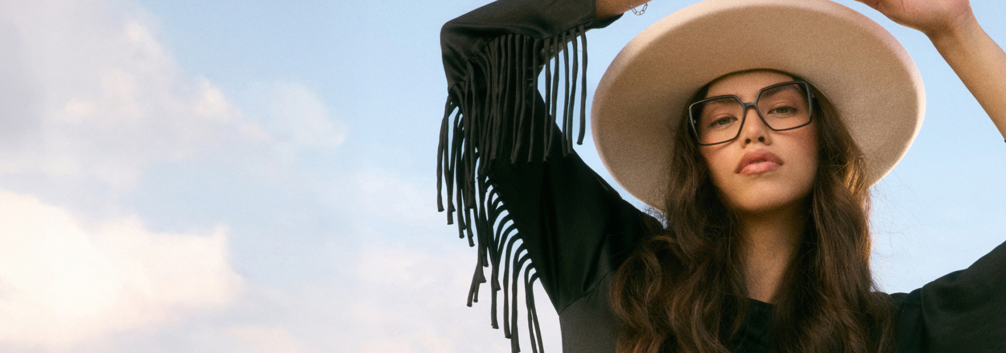 Person wearing a wide-brimmed hat and glasses, with a fringed sleeve, against a blue sky.