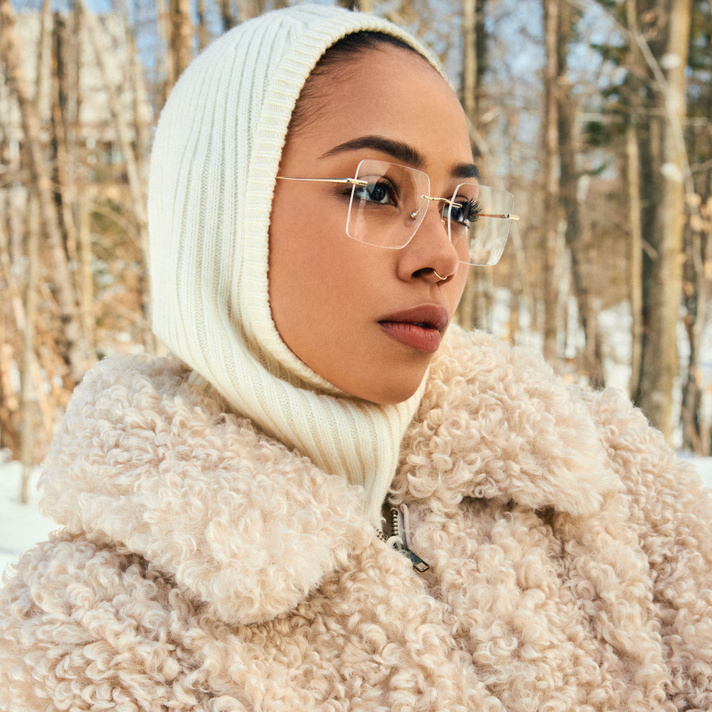 Person in a fluffy coat and glasses with a snowy forest background.