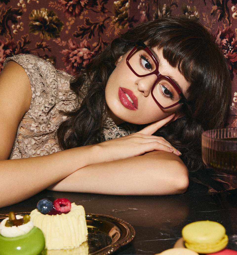 A woman in red glasses rests her head on her arms at a table with small cakes.