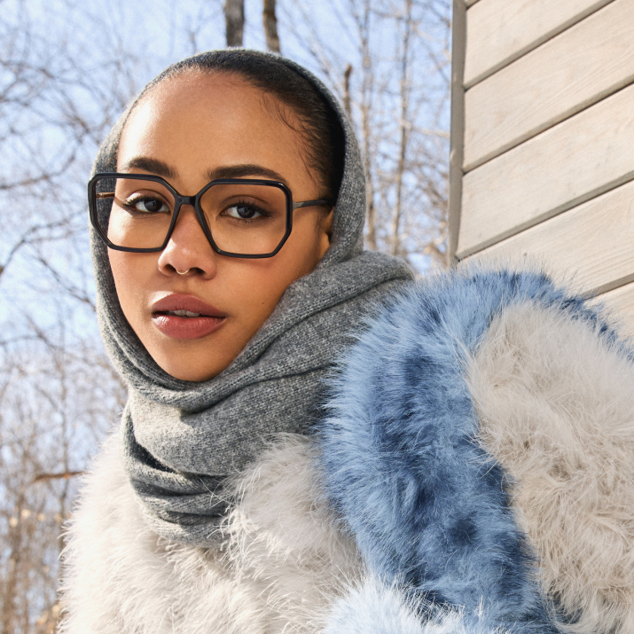 Person wearing glasses and a fur-trimmed coat, with trees in the background.