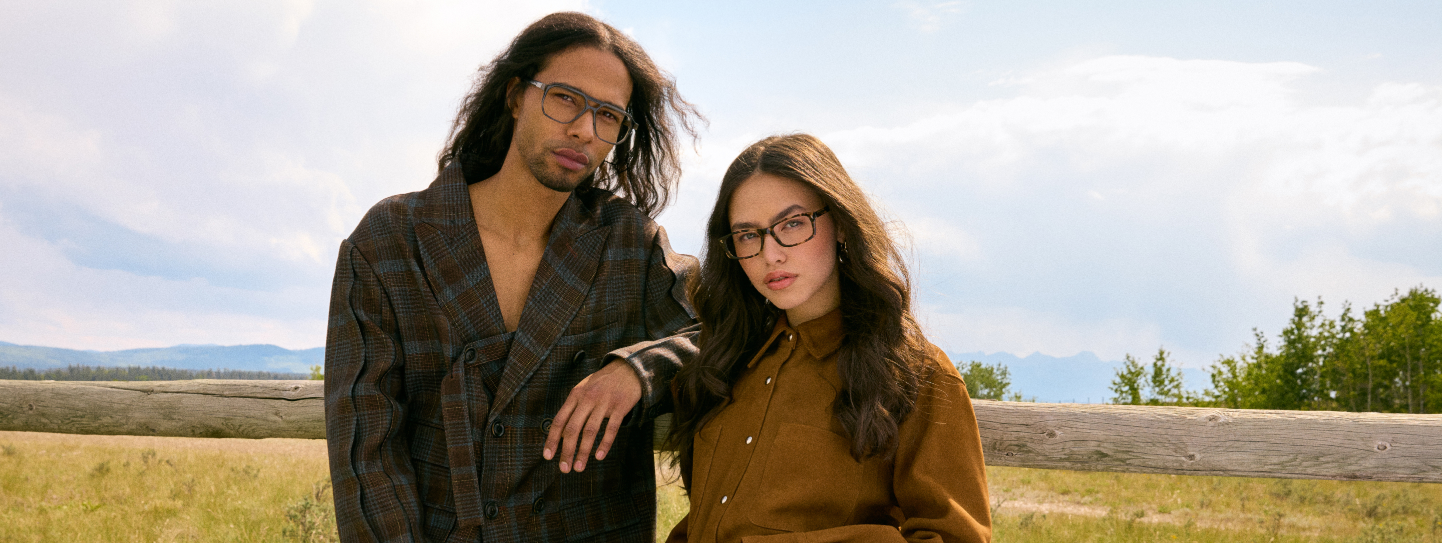 Two people wearing glasses standing by a wooden fence in a rural landscape.