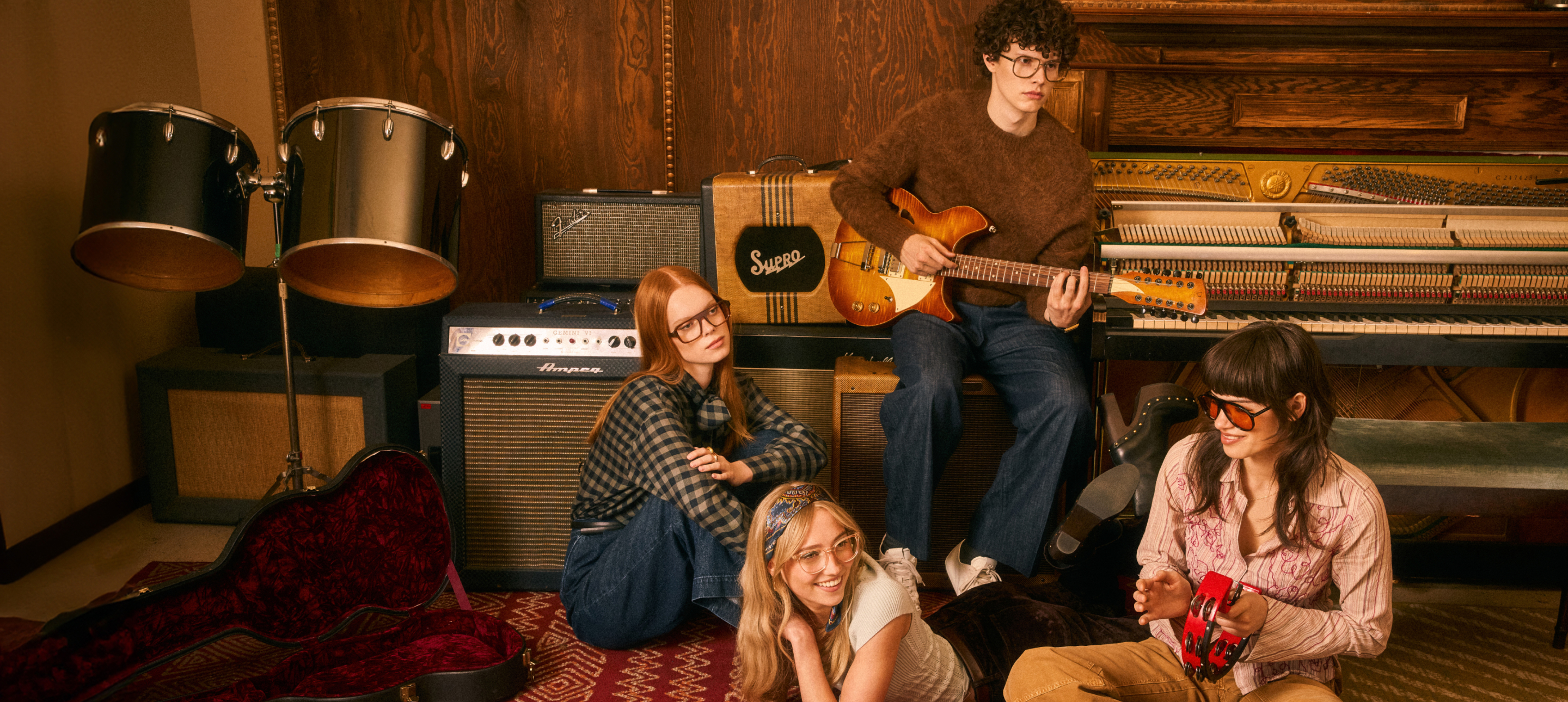 Four young musicians with vintage instruments and amplifiers in a wood-paneled room.