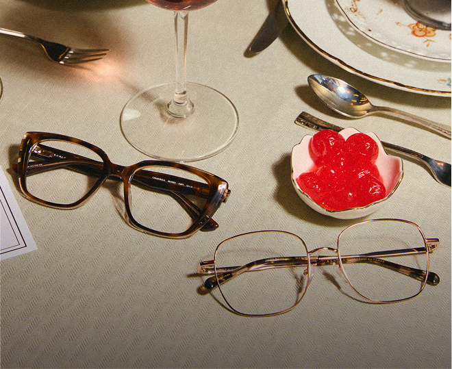 Two pairs of eyeglasses on a dinner table next to a glass of wine and red candies.