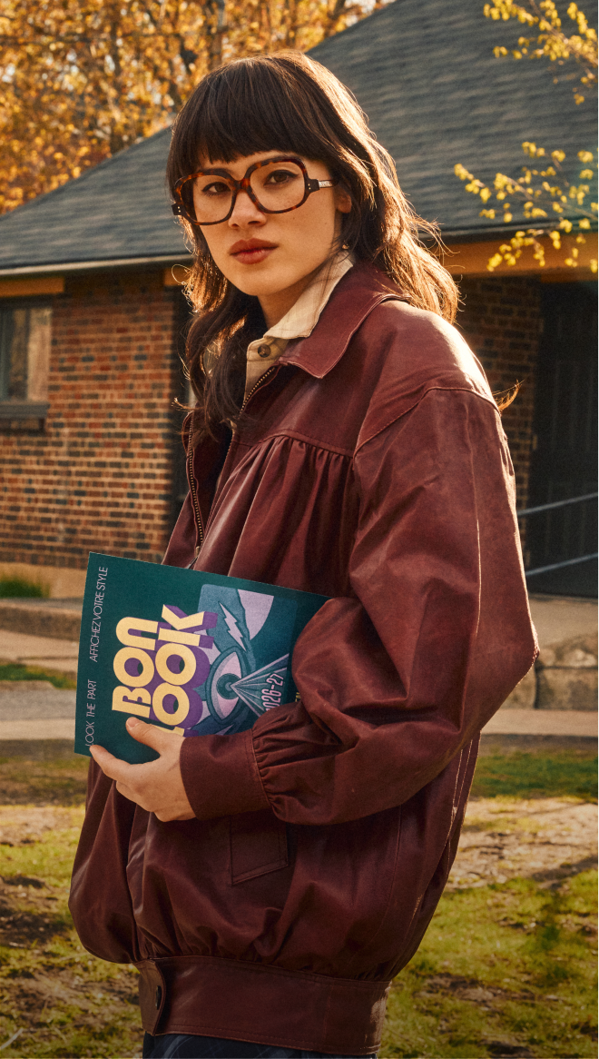A woman in tortoiseshell glasses and a reddish-brown jacket holds a book while standing outdoors.