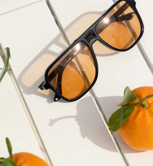 Sunglasses with orange lenses on a white surface, next to oranges.