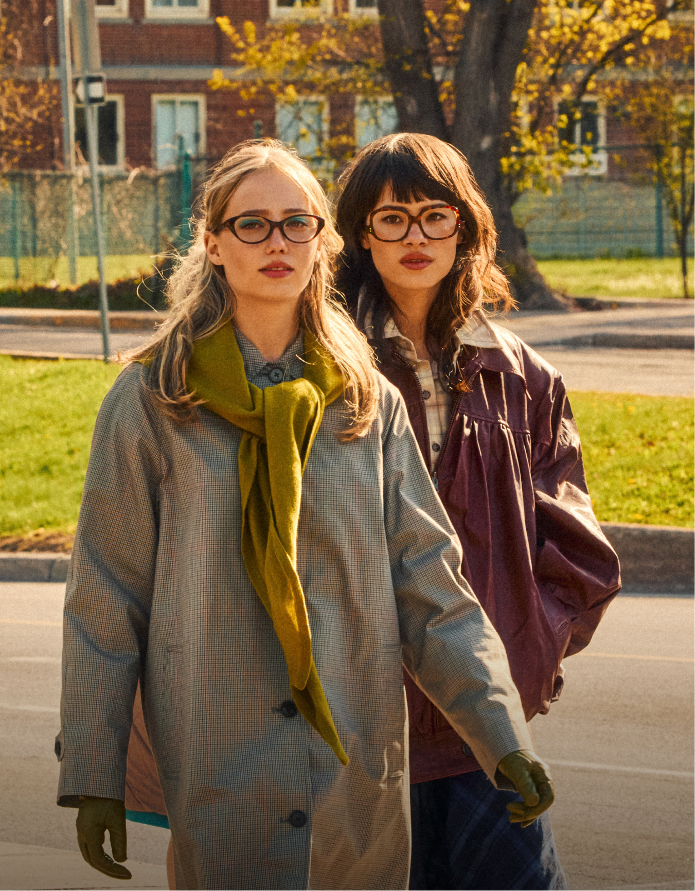 Two women wearing glasses and autumn coats stand side-by-side on a street during the day.