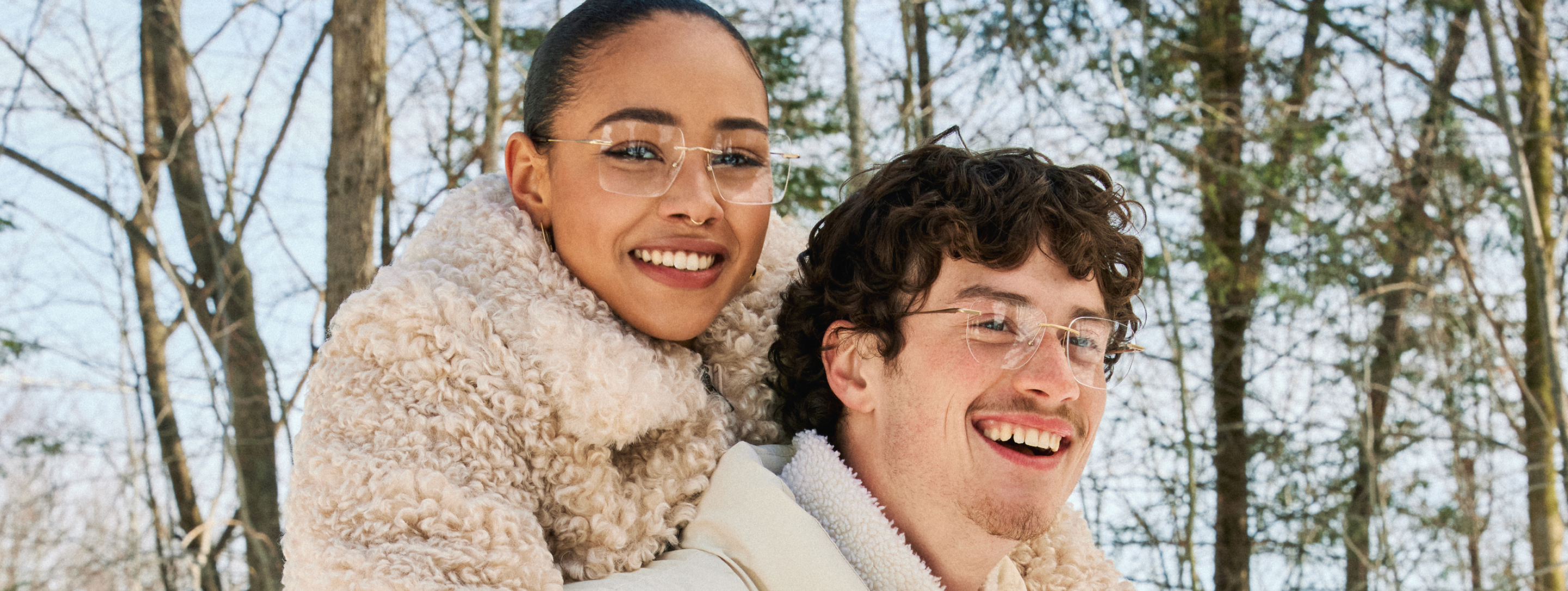 Two people smiling in a forest, wearing glasses and warm clothing.