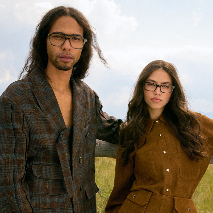 Two people wearing glasses and stylish outfits, standing outdoors with a cloudy sky background.