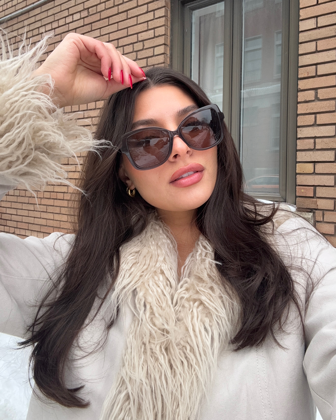 A woman with dark hair and sunglasses poses in a white coat with a shaggy collar.
