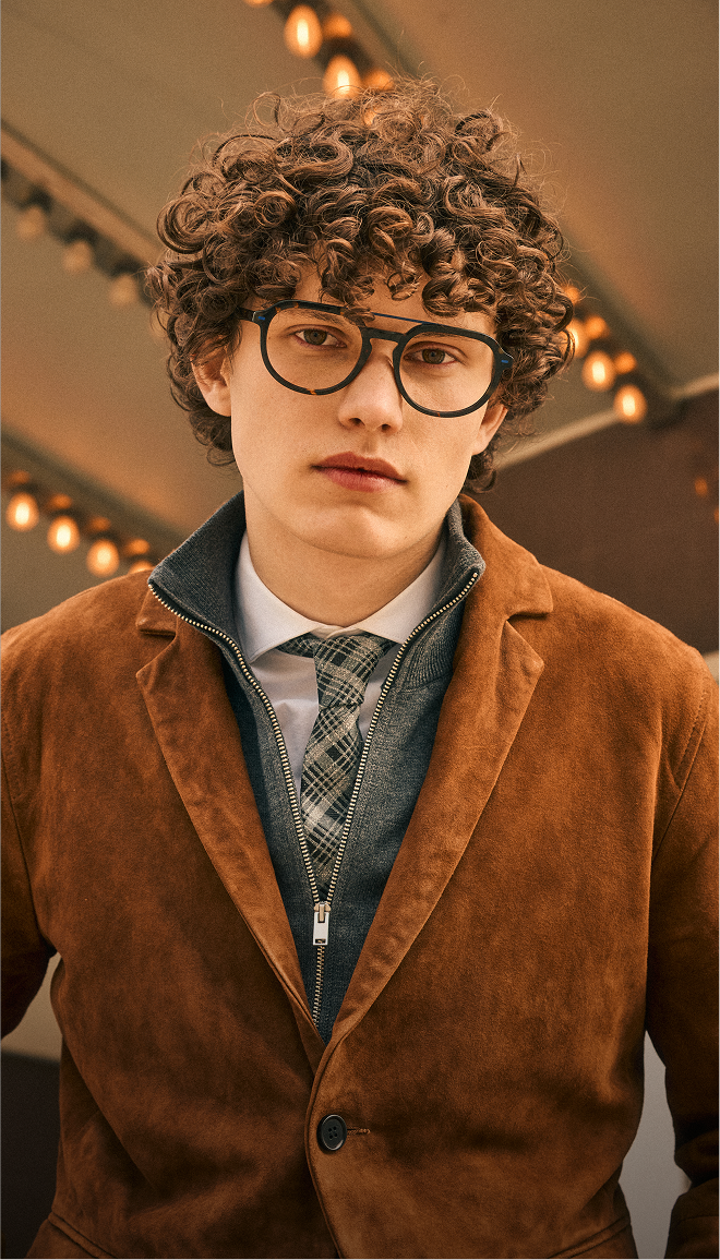 A young man with curly hair and glasses wearing a layered outfit with a brown suede jacket.