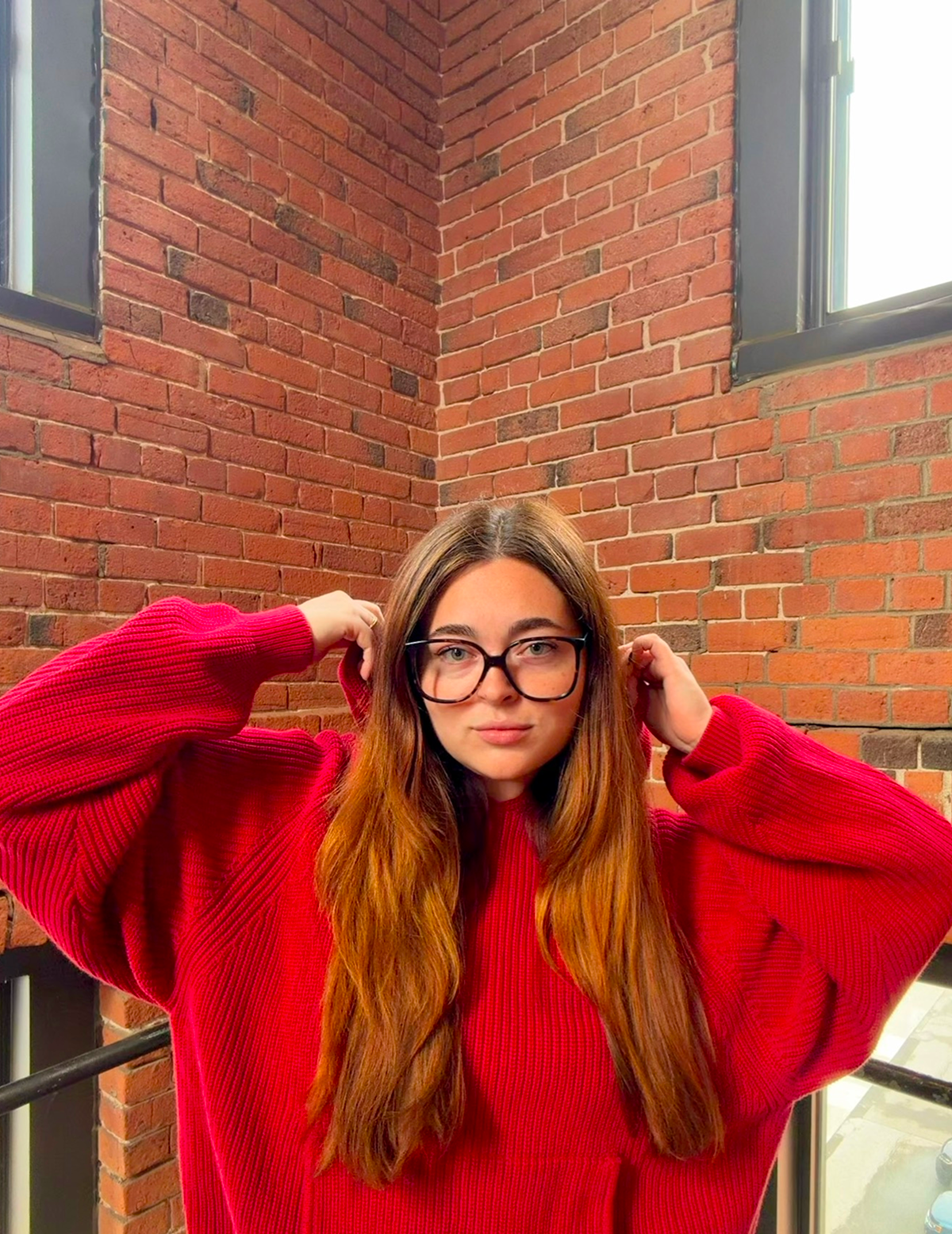 A young woman in glasses and a red knit hoodie stands against a brick wall.