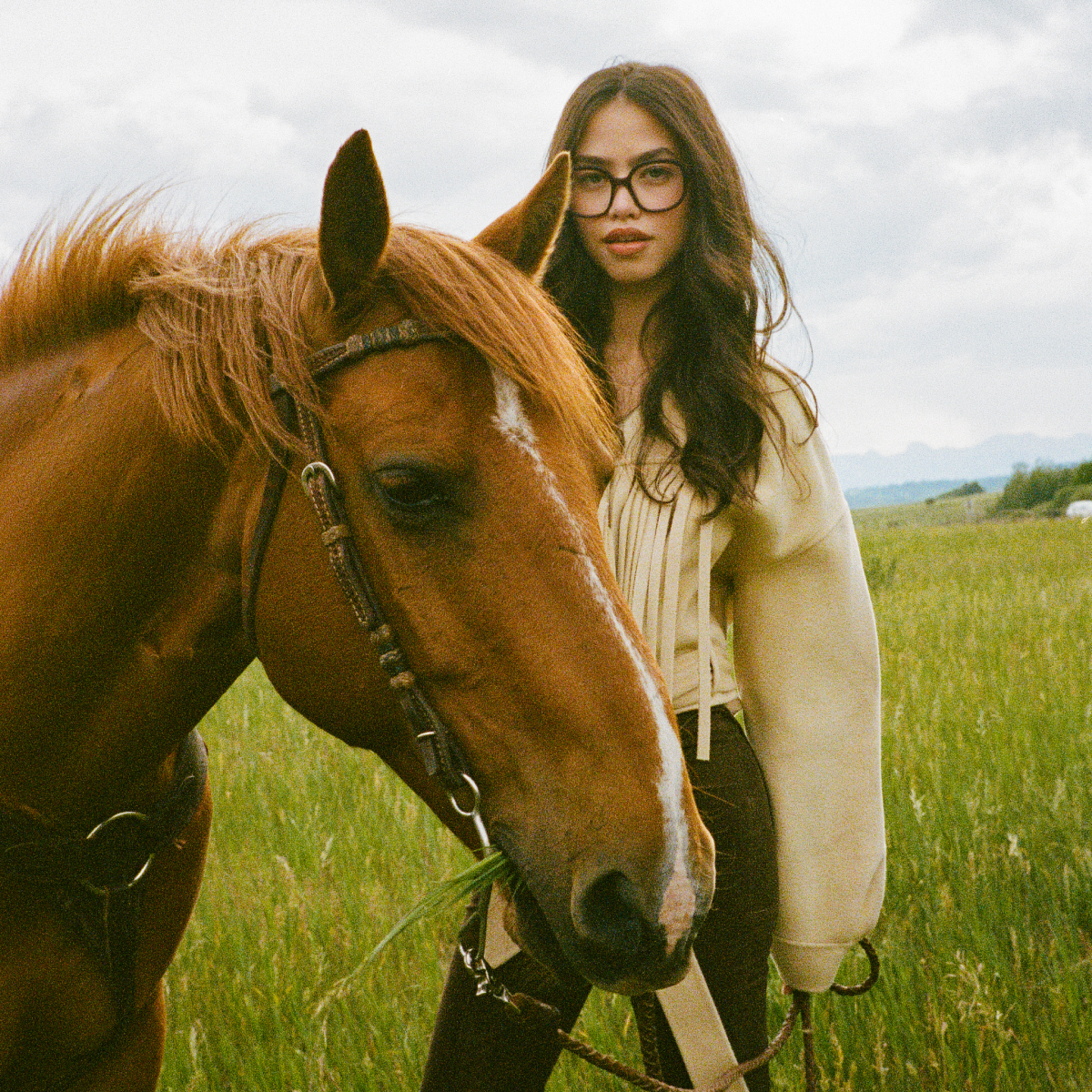Woman with glasses standing beside a brown horse in a grassy field.