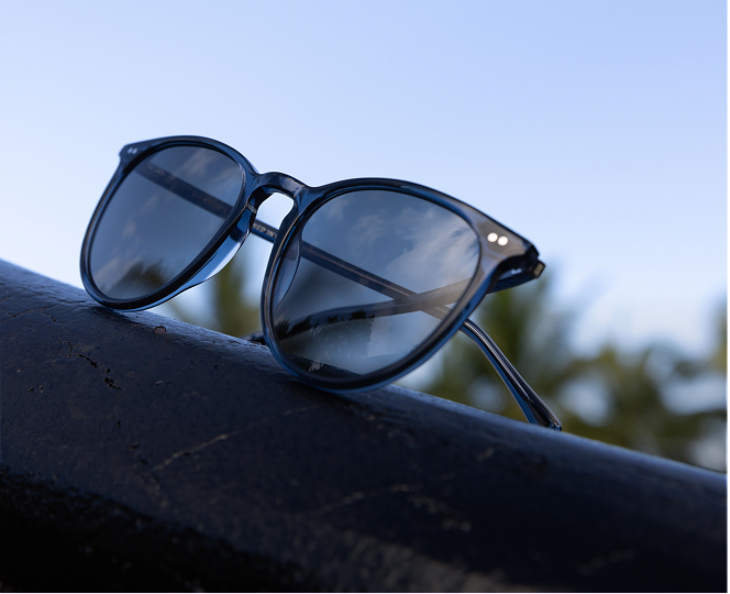 A pair of blue-framed sunglasses resting on a dark railing against a clear blue sky.